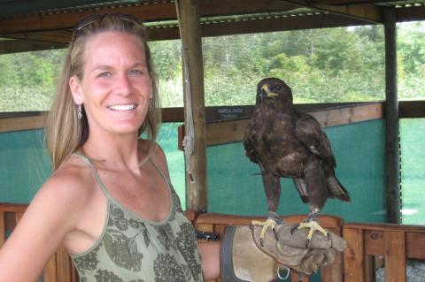 a smiling woman with long blond hair wearing a tank top. She has a steppe eagle perched on her arm.